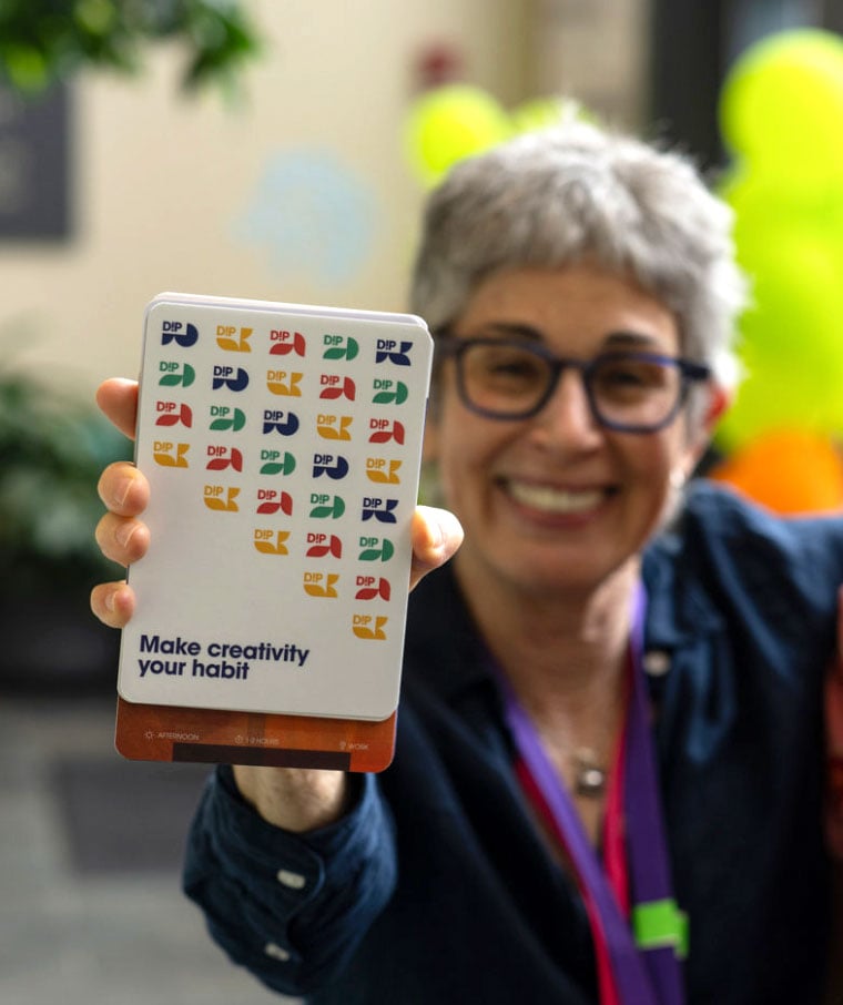 A woman raising a postcard to the camera that reads "Make creativity your habit"